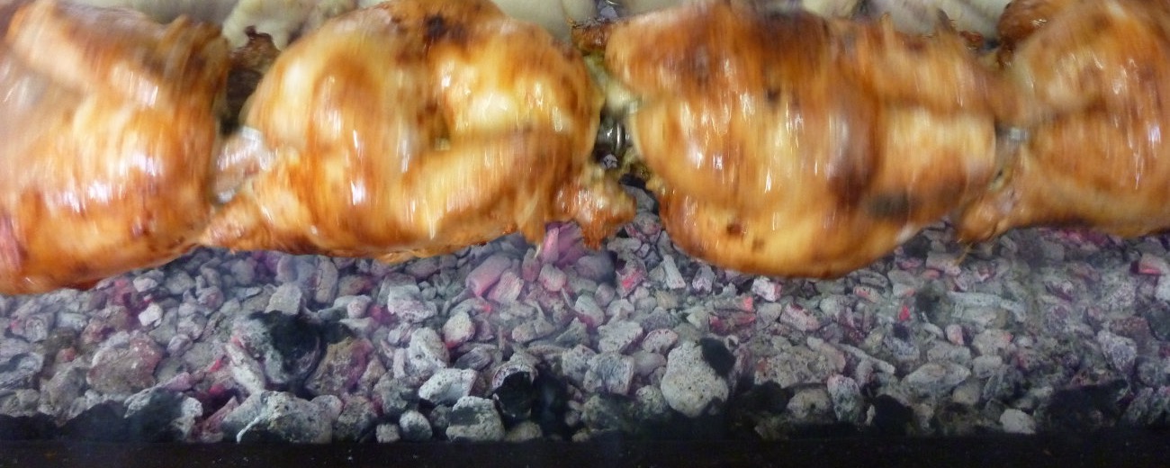 A row of golden charcoal chickens turning on the rotisserie over glowing coals