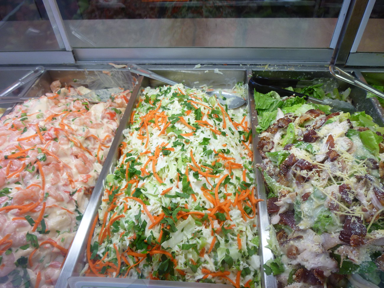Three trays in the salad bar: seafood salad, coleslaw, and chicken Caesar salad
